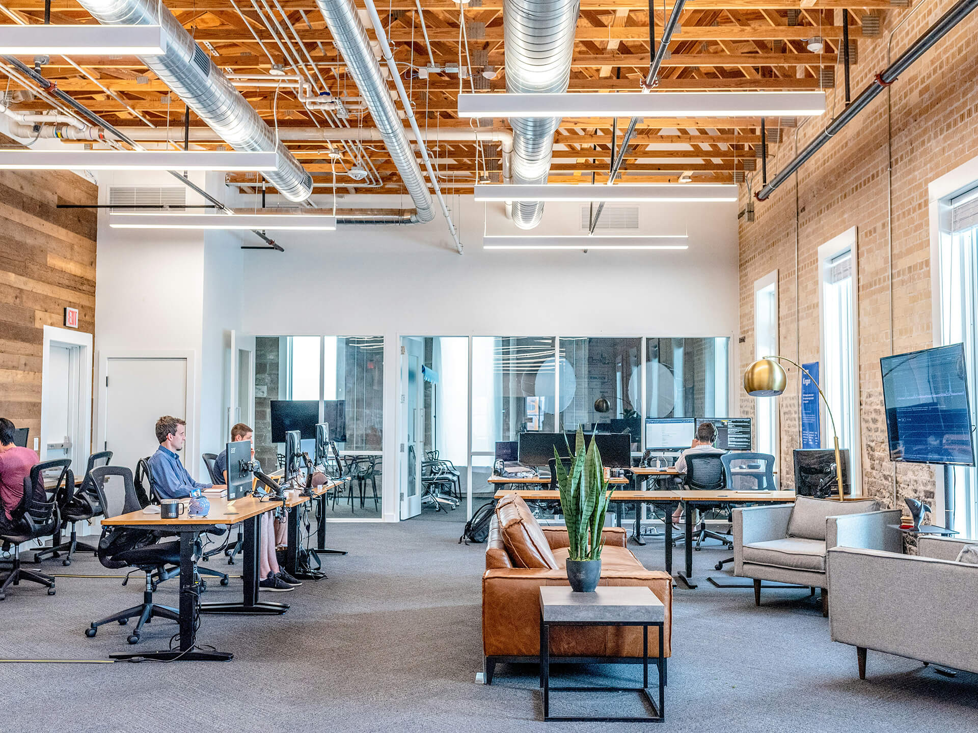 An open-concept office with high ceilings, warm wood and brick walls, and 4 people sitting at desks working on their computers.